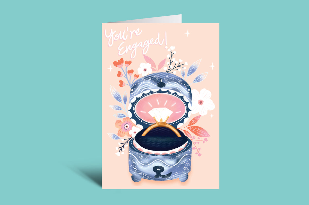 Engagement card with a blue ring box and floral design on a pink background