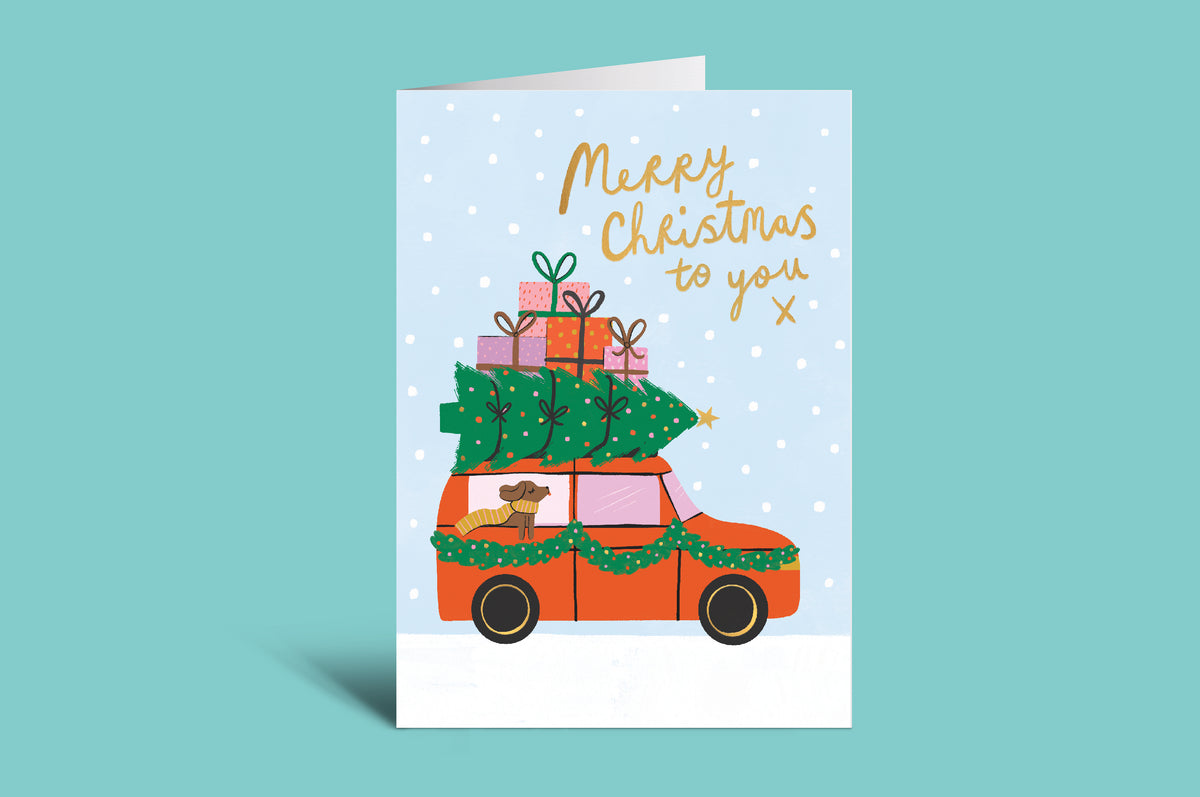 Christmas card with a car carrying a Christmas tree and presents on a light blue background.
