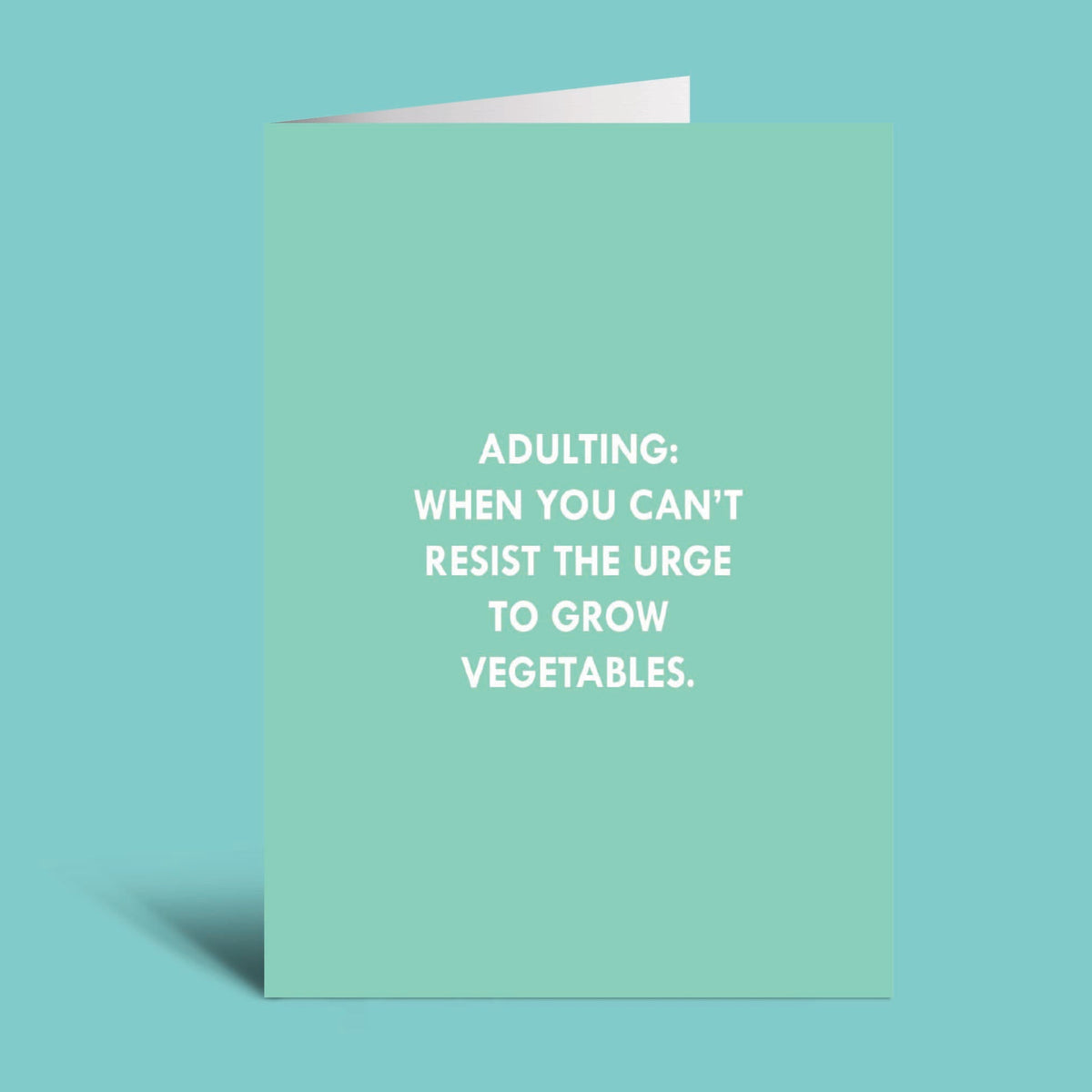 A mint green greeting card with white text that reads 'ADULTING: WHEN YOU CAN'T RESIST THE URGE TO GROW VEGETABLES.' shown against a solid teal background.