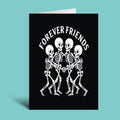 A black greeting card standing upright against a light aqua background, printed with the phrase 'FOREVER FRIENDS' above a white graphic of four skeletons holding hands.