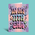A greeting card with retro 3D bubble lettering that reads 'IN MY 11TH ERA' in pastel rainbow colors, surrounded by musical notes and stars on a purple and pink sunburst background, shown standing against a solid teal backdrop.