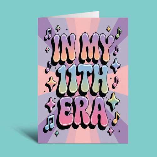 A greeting card with retro 3D bubble lettering that reads 'IN MY 11TH ERA' in pastel rainbow colors, surrounded by musical notes and stars on a purple and pink sunburst background, shown standing against a solid teal backdrop.