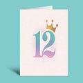 A folded greeting card standing upright on a teal background. The card is off-white with a large stylized number 12 in a pastel gradient of pink, purple, and blue, topped with a small gold crown illustration.