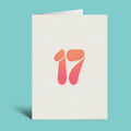 A folded greeting card standing upright on a light teal background. The card is off-white with a stylized number 17 printed in the center, featuring a warm orange-to-pink gradient.