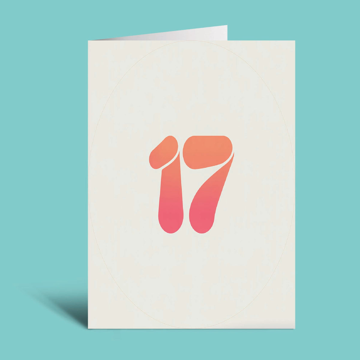 A folded greeting card standing upright on a light teal background. The card is off-white with a stylized number 17 printed in the center, featuring a warm orange-to-pink gradient.