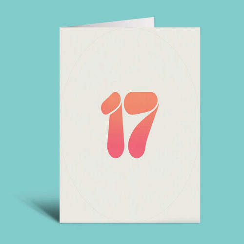 A folded greeting card standing upright on a light teal background. The card is off-white with a stylized number 17 printed in the center, featuring a warm orange-to-pink gradient.