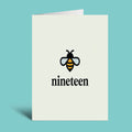 A vertical cream-colored greeting card standing upright on a light blue background. The card features a black and yellow bumblebee graphic above the word 'nineteen' in a black serif font.