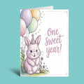 A children's first birthday greeting card featuring a cute watercolor-style white bunny wearing a party hat, surrounded by pastel balloons and small flowers, with the text 'One Sweet Year!' on a white card against a teal background.