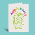 A folded greeting card standing upright on a light aqua background. The card is white with a colorful 'Happy 3rd Birthday' script and a lime green cartoon crocodile wearing sunglasses.