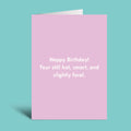 A pink greeting card with white text reading 'Happy Birthday! Your still hot, smart, and slightly feral.' shown standing against a teal background.