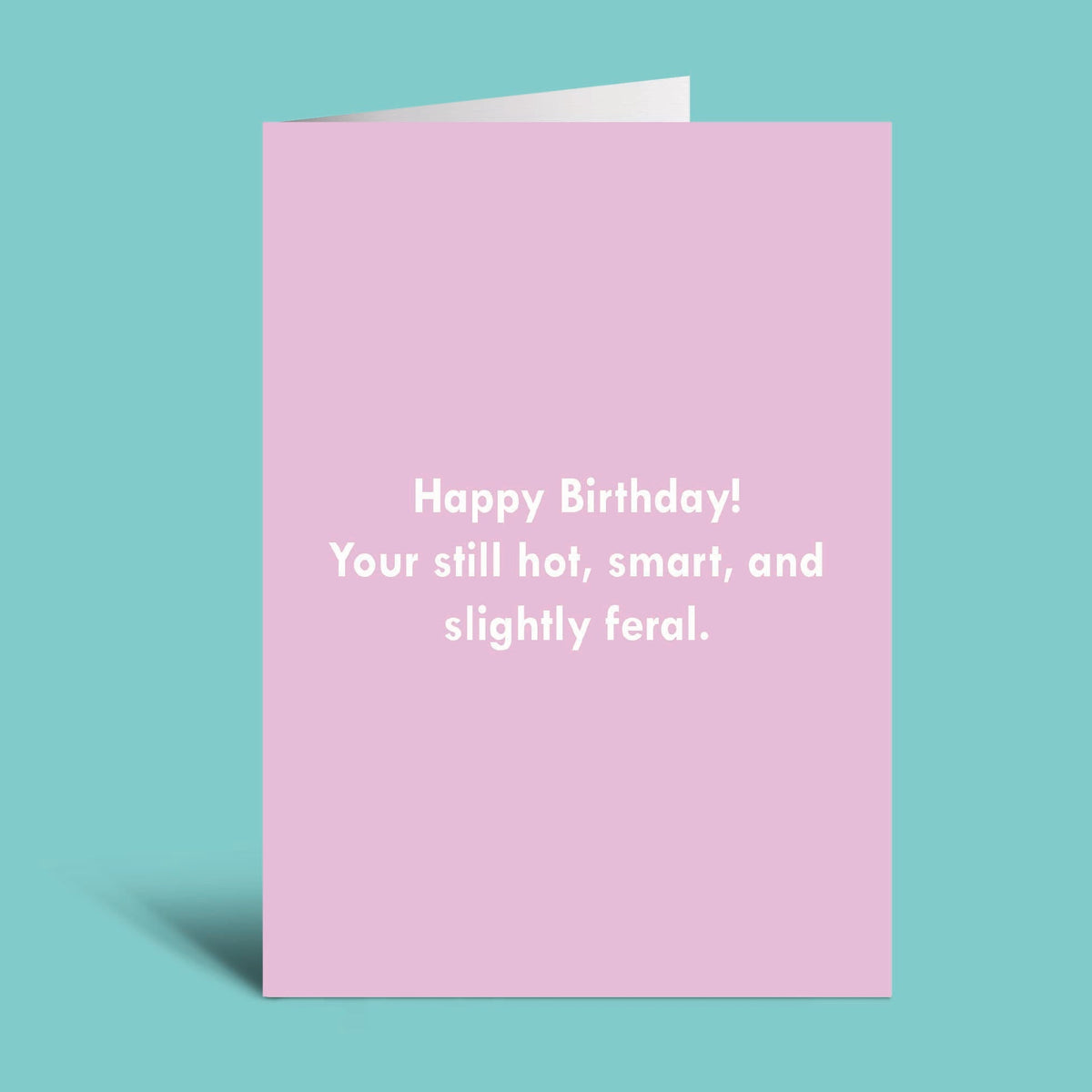 A pink greeting card with white text reading 'Happy Birthday! Your still hot, smart, and slightly feral.' shown standing against a teal background.