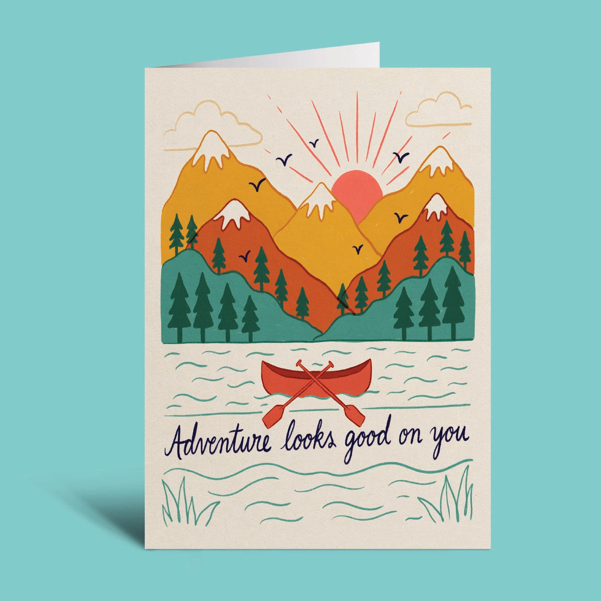 A vertical greeting card with a colorful illustrated mountain landscape, sun, trees, and a red canoe on the water, with the text 'Adventure looks good on you' at the bottom. The card is shown standing upright against a solid light aqua background.